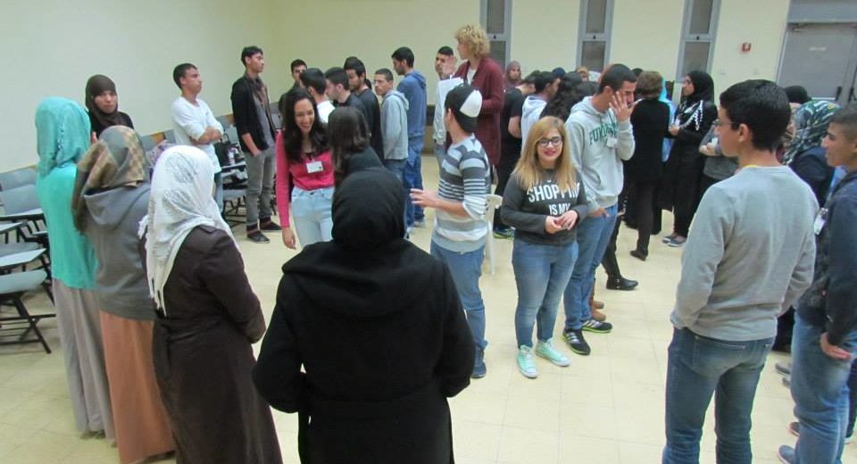 Sharón stands on a chair in the center of a circle of students, leading an introductory game at a Young Negotiators Training seminar with Arab and Jewish Israeli high school students.